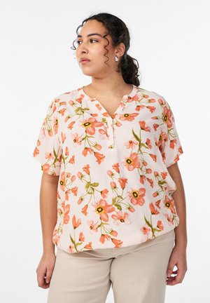 Femme aux cheveux bouclés et foncés attachés en queue de cheval, portant un chemisier blanc à manches courtes à motifs floraux et un pantalon beige, regardant sur le côté.