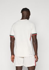 Maglietta atletica bianca con texture a coste, maniche corte con accenti rossi e verdi, e scollatura rotonda. Pantaloni corti dello stesso colore.