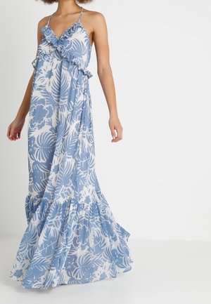 Femme portant une longue robe bleu et blanc à motifs floraux avec des volants, debout devant un fond blanc uni.