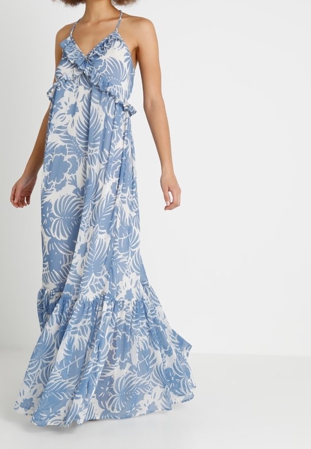 Femme portant une longue robe bleu et blanc à motifs floraux avec des volants, debout devant un fond blanc uni.