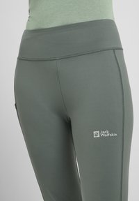 Donkergroene leggings van rekbaar materiaal met een gladde textuur. Heeft een brede tailleband en een logo op het linkeronderbeen.