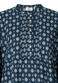 Blusa blu navy con motivo floreale a rombi bianchi, dotata di patta con bottoni, plissettature e una vestibilità ampia con un orlo inferiore arricciato.