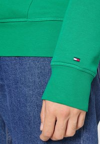 Primo piano di una mano appoggiata su jeans di denim blu, che indossa una manica di un felpa verde con un piccolo logo rosso, bianco e blu vicino al polsino.