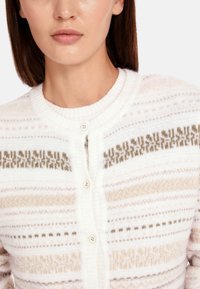 Weißer Strickcardigan mit horizontalen Streifen in Gold und Braun. Er hat einen runden Ausschnitt und fünf Knopfverschlüsse auf der Vorderseite.