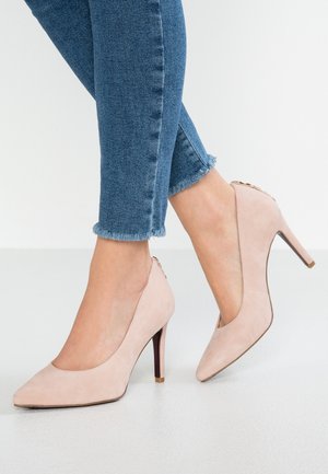 Person iført blå skinny jeans med flossede kanter og lyserøde højhælede sko står på et hvidt gulv.
