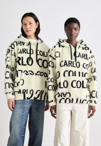 Fluffy creme hoodie med sort tekst "CARLO" og grafik. Løs pasform, frontlommer, snore, båret med jeans og lyse bukser.