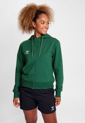 Groene rits hoodie van zacht materiaal, met een voorkantzak, trekkoorden en logodetail. Gecombineerd met zwarte shorts.