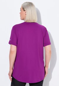 Blond kvinde med kort hår iført en løs lilla kortærmet skjorte, vendt væk, mod en ensfarvet hvid baggrund.