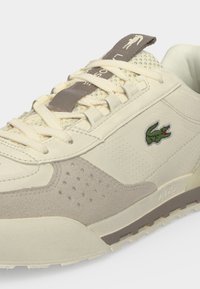 Μπεζ και γκρι sneaker Lacoste με κορδόνια, διάτρητες σουέντ λεπτομέρειες, γλωττίδα από δίχτυ και πράσινο λογότυπο κροκοδείλου στο πλάι.