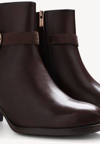 Bottes chelsea en cuir marron avec une finition brillante, fermeture éclair sur le côté, et une sangle en daim avec un ornement métallique circulaire.