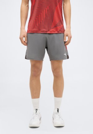 Čovjek u crvenoj sportskoj majici s uzorkom, sivim sportskim kratkim hlačama, bijelim čarapama i bijelim tenisicama, stoji nasuprot jednostavne pozadine.