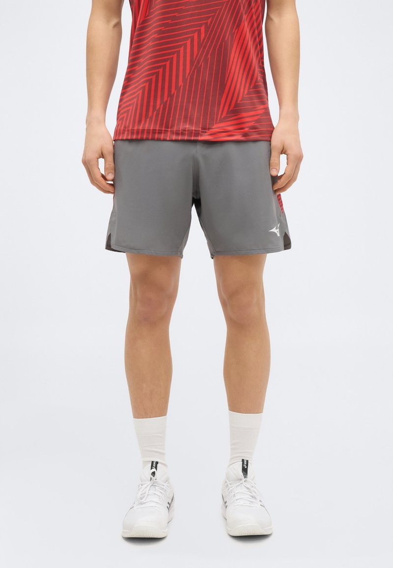 Άνδρας που φοράει κόκκινο αθλητικό πουκάμισο με σχέδια, γκρι αθλητικό σορτς, λευκές κάλτσες και λευκά αθλητικά παπούτσια, στέκεται μπροστά σε απλό φόντο.