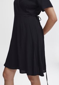 Robe noire cache-coeur en tissu doux, dotée d'un lien à la taille et de manches courtes, avec un ourlet évasé atteignant le mi-thigh.