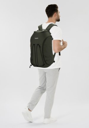 Uomo che indossa una maglietta bianca, pantaloni grigio chiaro e sneaker bianche, mentre porta uno zaino grande di colore verde scuro su uno sfondo bianco semplice.