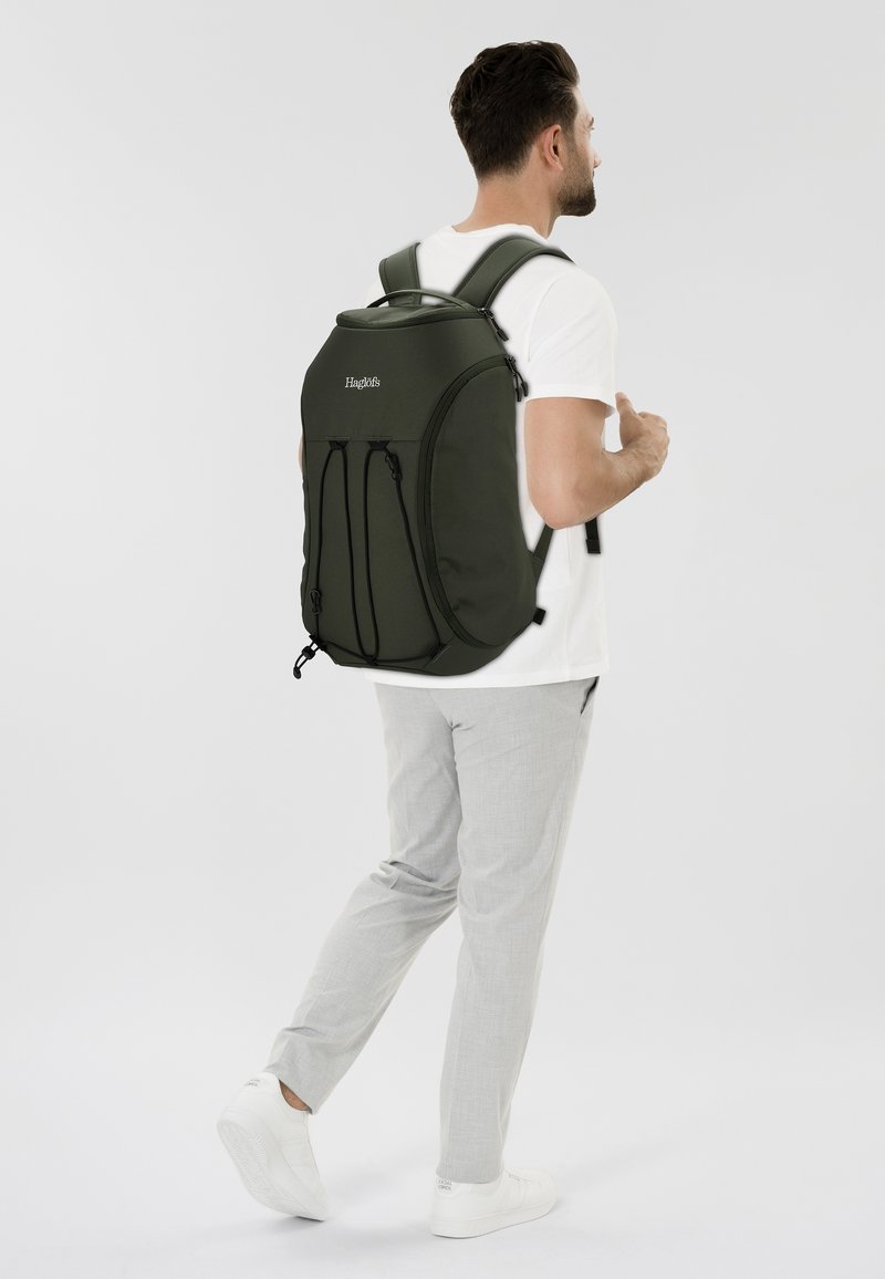 Man met een wit T-shirt, lichtgrijze broek en witte sneakers, die een grote donkergroene rugzak draagt tegen een effen witte achtergrond.