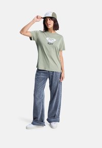 Olivově zelené tričko s bílým potiskem loga, v kombinaci s širokými šedými džíny a bílými teniskami. Na hlavě má čepici s zelenými a bílými akcenty.