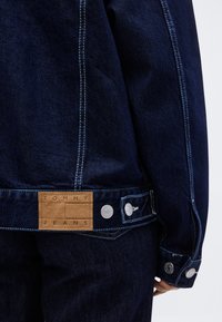 Σκούρο μπλε τζιν μπουφάν με λευκή ραφή, καφέ δερμάτινη ετικέτα "Tommy Jeans" και ασημένια μεταλλικά κουμπιά στη μανσέτα και τη μέση.