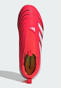 Botín de fútbol rojo con un upper suave y texturizado, con distintivas rayas en negro y blanco, y una lengüeta en la parte trasera para facilitar su uso.