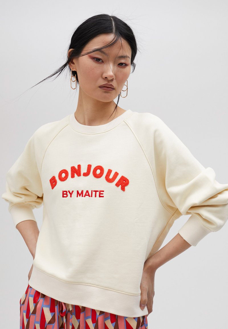 Felpa crema con ricamo rosso "BONJOUR BY MAITE", maniche raglan e polsini a coste, abbinata a pantaloni a fantasia.