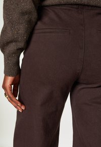 La mano di una persona con anelli d'oro posata di lato, che indossa pantaloni marroni scuri e una manica di un maglione marrone maculato visibile.