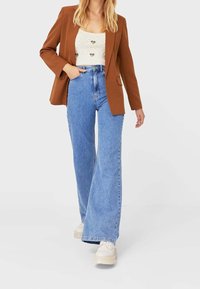 Blazer marron sur un haut en tricot léger, associé à un jean bleu taille haute et large, et des chaussures à plateforme. Tenue simple et décontractée.
