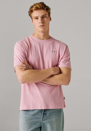 Młody mężczyzna z założonymi skrzyżowanymi rękami, ubrany w jasnoróżową koszulę z krótkim rękawem i jasnoniebieskie dżinsy na jednolitym szarym tle.