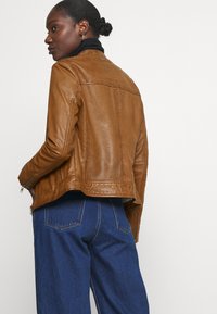 Brown leather jacket with a fitted design, featuring zippered cuffs and stitching details, worn over a black turtleneck. Dark blue jeans.