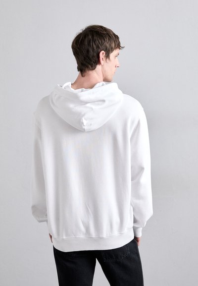 Felpa bianca con un ampio cappuccio, polsini e orlo a coste. Tessuto morbido con una vestibilità comoda. Indossata con pantaloni neri, vista da dietro.
