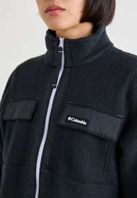 Czarna kurtka polarowa z wysokim kołnierzem, zamkiem z przodu i dwoma kieszeniami na piersi. Wykonana z tkaniny o fakturze oraz z kontrastowych, gładkich paneli.