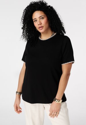 Femme aux cheveux bouclés portant un haut noir à manches courtes avec bordure blanche et un pantalon crème, accessoirisée avec des bracelets et des bagues.