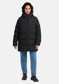 Giacca a vento nera con cappuccio, dotata di chiusura con zip e trapuntatura orizzontale. Abbinata a jeans azzurri chiari e calzature nere.