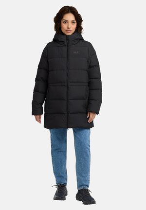Zwarte pufferjas met een capuchon, voorzien van een ritssluiting en horizontale quiltstructuur. Gecombineerd met lichtblauwe jeans en zwarte schoeisel.
