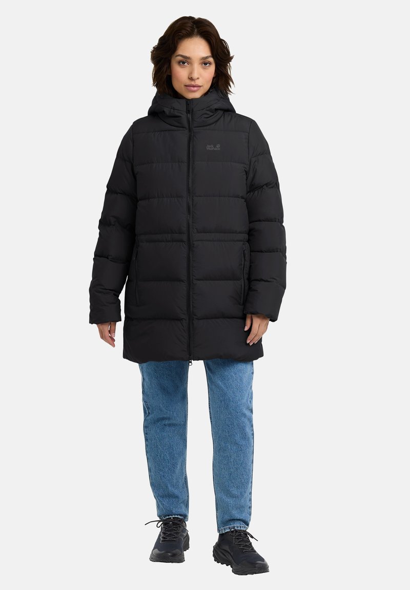 Giacca a vento nera con cappuccio, dotata di chiusura con zip e trapuntatura orizzontale. Abbinata a jeans azzurri chiari e calzature nere.