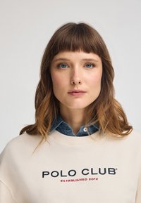 Maglione leggero beige con il logo "POLO CLUB" in blu e rosso, indossato sopra una camicia di jeans blu, con scollatura rotonda e texture morbida.