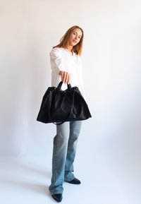 Mujer con camisa blanca y vaqueros azules sostiene un gran bolso negro hacia adelante sobre un fondo blanco liso.