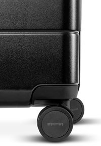 Zwarte harde koffer met een gestructureerd oppervlak, voorzien van vier duurzame wielen, ronde hoeken en gegraveerde branding op één wiel.