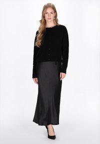 Cardigan nero con chiusura a bottoni, tessuto a maglia testurizzato, abbinato a una gonna di raso nera lunga fino al pavimento. Il modello indossa scarpe con tacco nere, capelli sciolti.