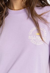Kvinna i en lavendelfärgad t-shirt med ett guldhalsband, prydd med en gul sol och vit text "SUNSHINE LOVERS BILLABONG ENDLESS."