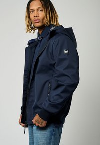 Veste bleu marine avec une fermeture éclair à l'avant, capuche ajustable, poignets élastiques et poches latérales. Présente un logo sur la manche gauche et un tissu texturé.