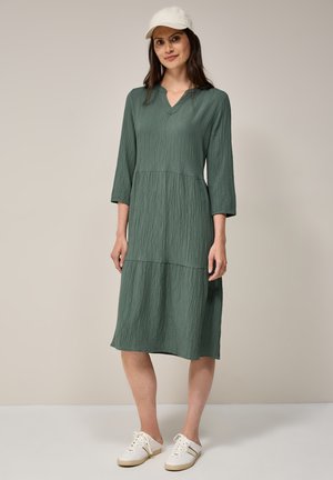 Vestido verde con textura, cuello en V, mangas tres cuartos y diseño en capas, combinado con zapatillas blancas y una gorra ligera.