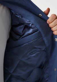 Navy outer fabric jacket with a quilted satin lining, featuring a side pocket and snap closure, showcasing smooth texture and rich color contrast.