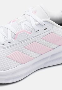 White athletic shoe with mesh upper, pink accents, and three stripes. Features textured detailing and traditional lace closure.