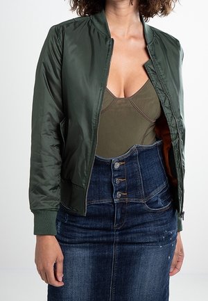 Olivově zelená bomber bunda s žebrovaným manžetovým výstřihem, zapínáním na zip a bočními kapsami, v kombinaci s upnutým béžovým body a modrou džínovou sukní s knoflíky.