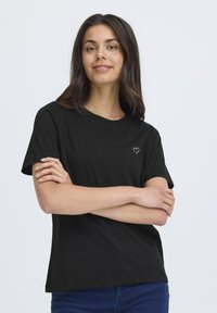 Juodas medvilnės marškinėliai trumpomis rankovėmis ir apvaliu iškirpimu, su maža balta širdelės dizaino detalė kairėje krūtinės pusėje.