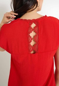 Blouse rouge avec un col rond orné de dentelle et un design dos nu avec un motif de dentelle florale. Tissu léger à texture.