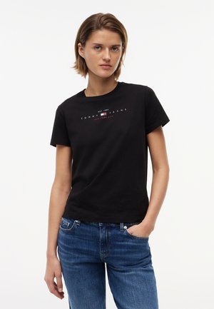 Mladá žena oblečená v čiernom tričku Tommy Jeans a modrých džínsoch, stojaca s jednou rukou v vrecku oproti bielemu pozadiu.