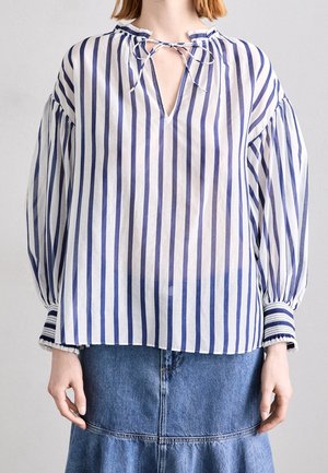 Kvinde iført en langærmet hvid bluse med vertikale marineblå striber, knyttet halsudskæring, og en blå denimnederdel.