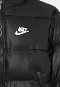 Svart polstret jakke med synlig glidelås og hvit Nike-logo på brystet, som viser detaljert søm og lommeomriss.