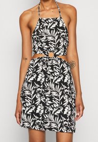 Schwarz-weißer Haltertop-Kleid mit einem Blattmuster, ausgeschnittenem Taillenbereich mit einem kreisförmigen Akzent und strukturiertem Stoff.