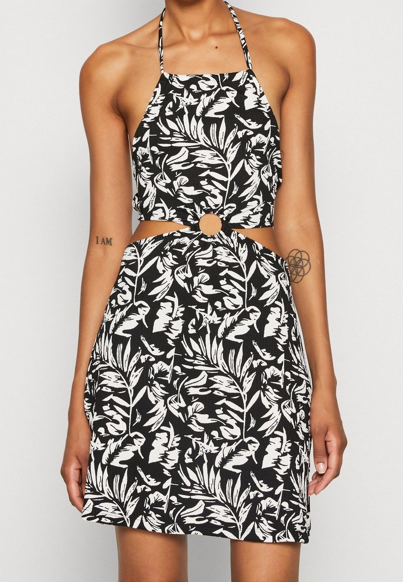 Schwarz-weißer Haltertop-Kleid mit einem Blattmuster, ausgeschnittenem Taillenbereich mit einem kreisförmigen Akzent und strukturiertem Stoff.